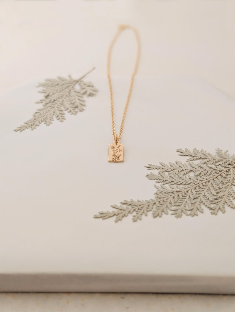 May include: A gold-colored necklace with a rectangular pendant featuring a floral design. The necklace is displayed on a white surface with decorative fern-like elements. The pendant is approximately 0.5 inches in length.