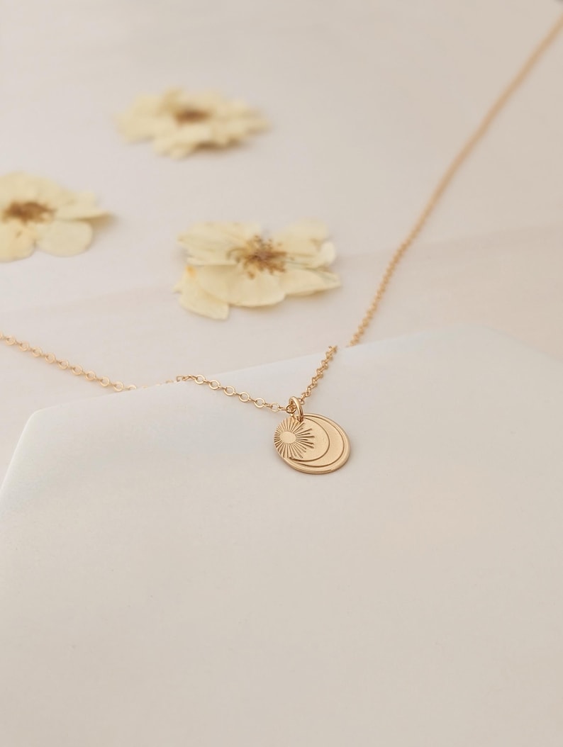 May include: A delicate gold-colored necklace featuring a pendant with sun and moon designs. The pendant hangs from a fine chain. The background includes dried flowers, creating a soft, natural aesthetic. The necklace is a piece of jewelry.