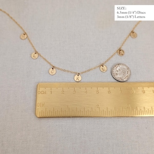 May include: Gold necklace with a series of small, round, gold charms with letters on them. The charms are hanging from a delicate gold chain. A ruler is included for size reference. The charms are 1/4 inch in diameter and the letters are 1/8 inch in diameter.