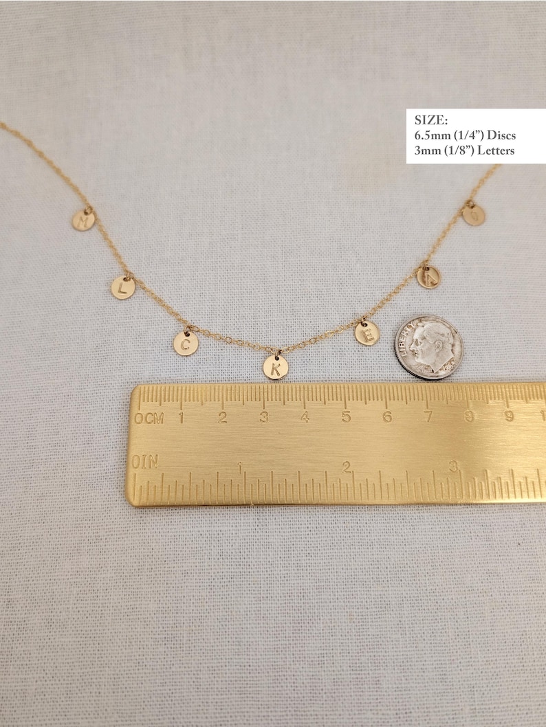 May include: Gold necklace with a series of small, round, gold charms with letters on them. The charms are hanging from a delicate gold chain. A ruler is included for size reference. The charms are 1/4 inch in diameter and the letters are 1/8 inch in diameter.