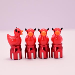 May include: Four red decorative valve stem caps. One is a duck, and the other three are bull-shaped. Each cap is mounted on a red and silver base. The duck has a black dot for an eye and an orange beak. The bull caps have black horns and orange beaks.