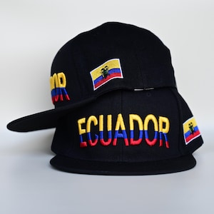 May include: Two black baseball caps with the word "ECUADOR" embroidered in yellow, blue, and red. Each cap features a small embroidered flag of Ecuador. The caps are stacked on top of each other.