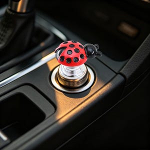 May include: A car accessory featuring a red ladybug with black spots and a black head, mounted on a circular silver-toned metallic base. The object is inserted into a vehicle's cigarette lighter socket, positioned on a black plastic center console.