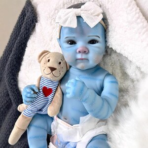 May include: A blue-skinned baby doll with a white bow headband and a plush teddy bear. The doll has detailed facial features and is wearing a diaper. The teddy bear has a blue and white striped shirt with a red heart.