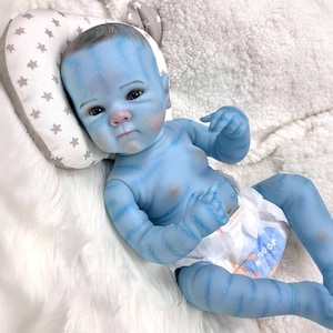 May include: A blue baby doll with detailed skin markings and a diaper. The doll has a serious expression and is lying on a white fluffy surface next to a pillow with star patterns.
