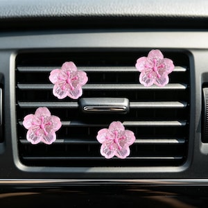 May include: Four pink, flower-shaped car air vent clips. Each clip has a translucent, petal design with a darker pink center. The clips are placed on a black car vent, adding a decorative touch to the vehicle's interior.