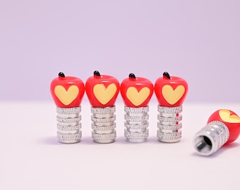 May include: Four red apple-shaped valve stem caps with yellow heart designs and black stems. The caps sit atop silver, threaded bases. One cap is lying on its side. The background is a soft purple.