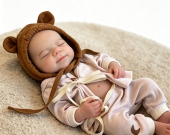 May include: A sleeping baby wearing a brown bear-eared hood and a light pink outfit. The outfit includes a jacket tied with a cream-colored ribbon and matching pants with a brown bear paw detail. The baby is lying on a beige rug.