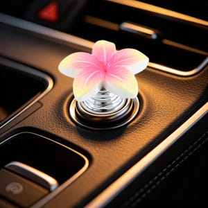 May include: A car interior accessory featuring a five-petaled flower with a gradient of pink at the center fading to white and yellow at the tips. The flower is mounted on a silver-colored circular base with a spring mechanism, attached to a dashboard.