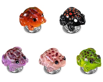 Könnte beinhalten: Fünf dekorative Schrankknöpfe in Froschform, jeder mit einer einzigartigen Farbgebung in Orange, Schwarz mit roten Punkten, Violett, Hellrosa und Grün mit dunklen Punkten, alle auf runden, silberfarbenen Metallsockeln montiert.