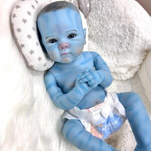 May include: A blue baby doll with detailed skin markings and a white diaper. The doll has a serene expression and is lying on a white fluffy surface, with a star-patterned pillow behind its head.