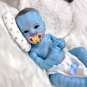 May include: A blue baby doll with detailed skin markings and a pacifier. The doll is lying on a white fluffy surface, with a small pillow with star patterns. The doll is wearing a diaper.