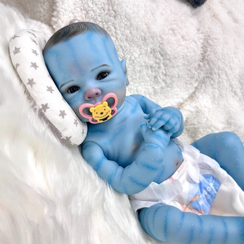 May include: A blue baby doll with detailed skin markings and a pacifier. The doll is lying on a white fluffy surface, with a small pillow with star patterns. The doll is wearing a diaper.