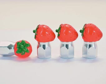 May include: A set of four silver-colored lug nuts, three topped with bright red strawberry-shaped caps with green stems. The fourth lug nut has a strawberry-shaped cap on its end. The background is plain white.