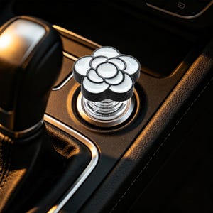 May include: A white and black flower-shaped car cigarette lighter plug insert. The accessory features a glossy white enamel-like finish on the petals with black outlines, set into a metallic silver base that fits into a standard vehicle socket.
