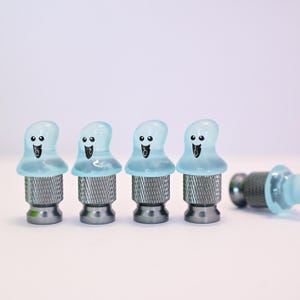 May include: Four blue ghost-shaped bottle stoppers with smiling faces and a silver base are arranged in a row. One stopper is lying on its side. The stoppers are made of glass and metal.