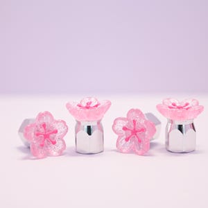 May include: Four decorative flower-shaped bottle stoppers. Each stopper features a pink, glittery flower design atop a silver-colored base. The background is a soft, light purple.