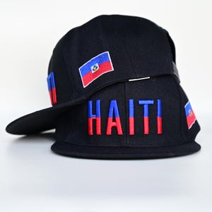 May include: Two black baseball caps stacked. The top cap has a small embroidered Haitian flag patch. The bottom cap features the word "HAITI" embroidered in blue and red. Additional flag patches are on the sides.