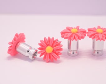 May include: Four daisy-shaped valve stem caps. The caps are pink with yellow centers and are mounted on silver metal stems. The caps are arranged on a light pink background.