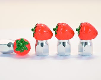 May include: A set of four silver-toned tire valve stem caps. Three caps are topped with bright red strawberry designs with green leaves. One cap is lying on its side, showing the strawberry design.