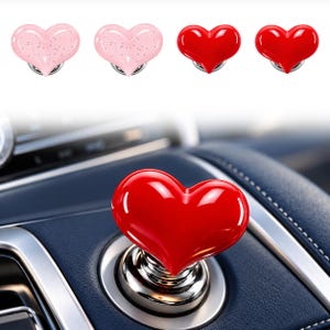 May include: A set of heart-shaped car interior accessories featuring two light pink glittery hearts and two solid red hearts. One red heart is shown installed on a circular silver-toned car dashboard button, while the others are displayed above.