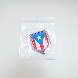 May include: A silver-colored shield-shaped emblem with a red, white, and blue Puerto Rican flag design. The flag features a white star on a blue triangle.