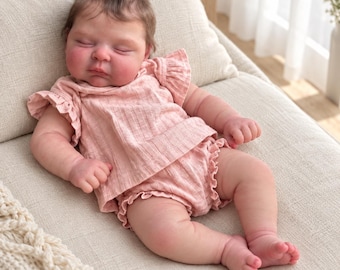 May include: A realistic newborn baby doll with closed eyes and soft brown hair, wearing a light pink ribbed two-piece outfit consisting of a short-sleeved top with ruffled sleeves and matching ruffled bloomers, lying in a relaxed sleeping position.