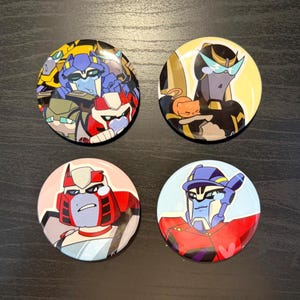Può includere: Quattro bottoni rotondi con spille che rappresentano personaggi di robot dei cartoni animati. I bottoni sono colorati e presentano una varietà di pose ed espressioni. I personaggi indossano tutti vestiti e accessori di diversi colori.