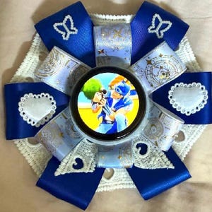 Könnte beinhalten: Eine handgefertigte Rosette mit einem zentralen Bild in einem schwarzen Kreis, umgeben von blauen und weißen Bändern. Das Design umfasst weiße Spitze, Schmetterlings- und Herzakzente sowie goldfarbene Verzierungen.