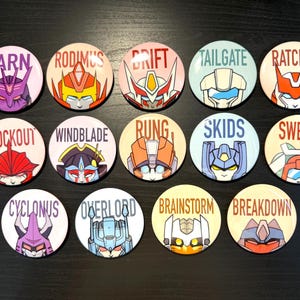 May include: Twelve round buttons featuring Transformers characters. Each button has a different character with their name printed above. The characters include Tarn, Rodimus, Drift, Tailgate, Ratchet, Knockout, Windblade, Rung, Skids, Swerve, Cyclonus, Overlord, Brainstorm, and Breakdown.