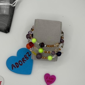 Könnte beinhalten: Vier farbige Perlenarmbänder mit verschiedenen Perlen in den Farben Grün, Orange, Rot, Lila und Braun. Die Armbänder werden auf einem grauen Samttuch präsentiert. Das Wort "ADORBZ" ist auf einem blauen herzförmigen Objekt geschrieben.