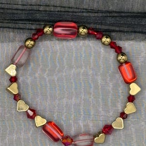 May include: A red and gold beaded bracelet with heart-shaped charms. The bracelet is made with red and clear glass beads, gold metal beads, and gold heart-shaped charms.