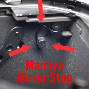 May include: Close-up of the mirror stop mechanism inside a Mamiya camera. The mechanism is made of black metal and has a small, black lever that is used to stop the mirror from flipping up.