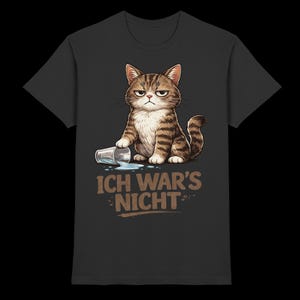 May include: A charcoal gray t-shirt featuring a graphic of a brown tabby cat with a grumpy expression. The cat has knocked over a clear glass, spilling liquid. Below the cat, the text reads "ICH WAR'S NICHT" in a bold, light brown font.