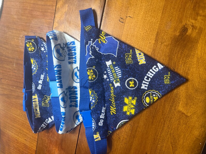 May include: Three triangular pet bandanas with blue adjustable straps and plastic buckles. Two bandanas feature a dark blue fabric with yellow and white Michigan team graphics, while one features a white fabric with blue Detroit Lions logos.
