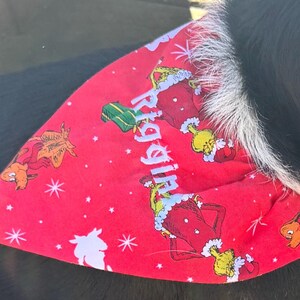 K&ouml;nnte beinhalten: Ein rotes, dreieckiges Haustierhalstuch mit einem wiederkehrenden Muster aus dem Grinch im Weihnachtsmannkost&uuml;m, gr&uuml;nen Geschenken und wei&szlig;en Sternen. Der Name Riggin ist in hellblauen Buchstaben diagonal aufgedruckt.