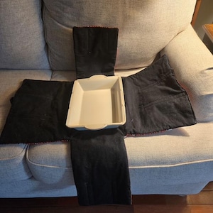 May include: A rectangular off-white ceramic baking dish centered on a black fabric carrier. The carrier features four extended flaps designed to fold over the dish, with visible stitching and pockets on the interior fabric panels.