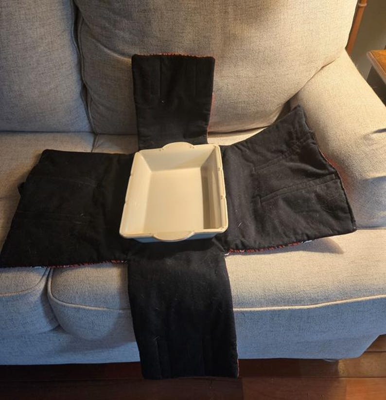 May include: A rectangular off-white ceramic baking dish centered on a black fabric carrier. The carrier features four extended flaps designed to fold over the dish, with visible stitching and pockets on the interior fabric panels.