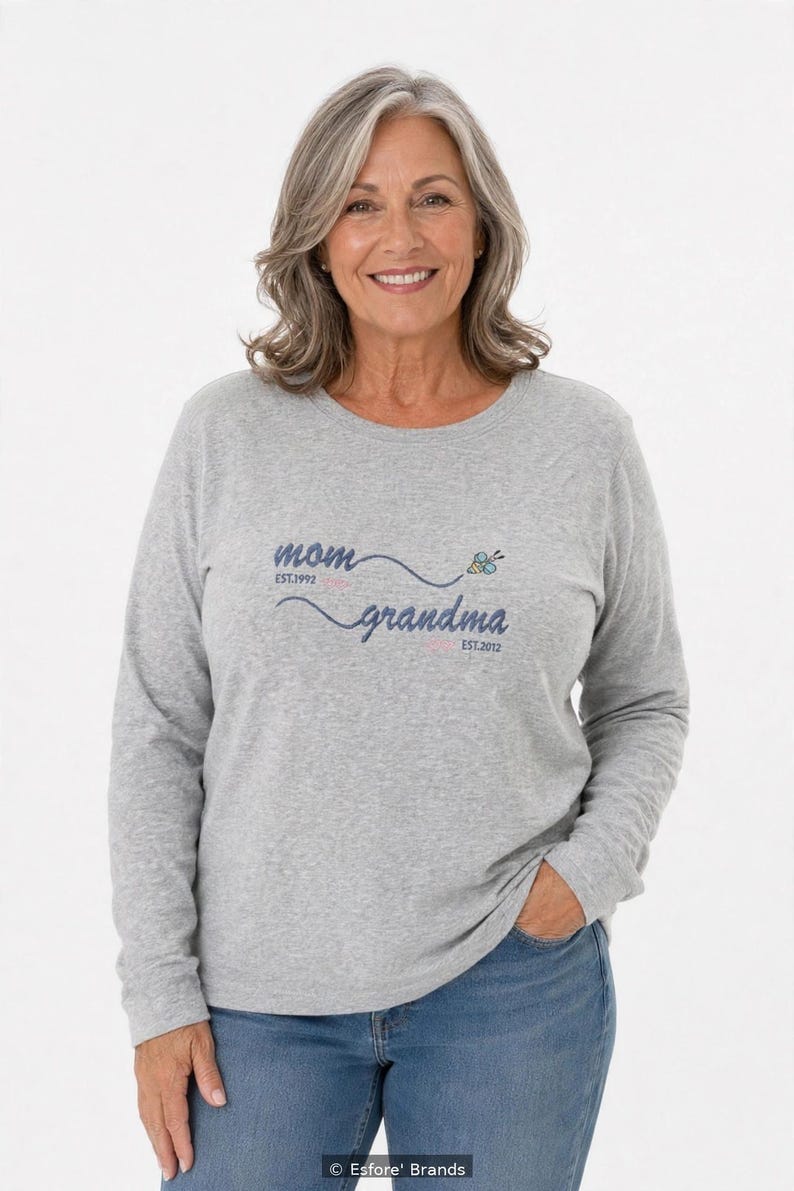 May include: A heather gray long-sleeve shirt featuring the text mom EST.1992 and grandma EST.2012 in dark blue script, connected by a wavy line with a small bee graphic. The shirt has a crew neckline and a relaxed fit, worn with blue jeans.