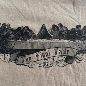 May include: A beige t-shirt featuring a black graphic print of a group of figures seated at a long table. Below the table, a banner displays the text The Final Table, with the logo for Fogo House Playing Cards printed underneath.