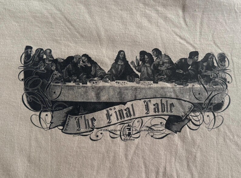 May include: A beige t-shirt featuring a black graphic print of a group of figures seated at a long table. Below the table, a banner displays the text The Final Table, with the logo for Fogo House Playing Cards printed underneath.