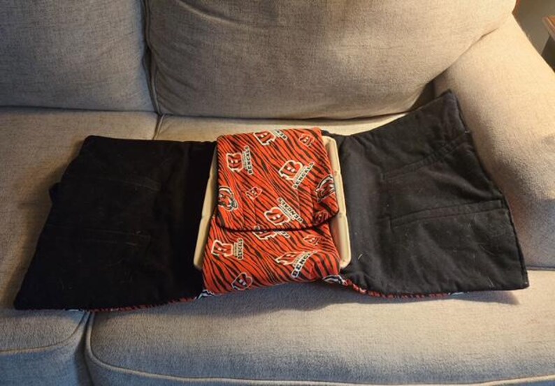 May include: A fabric lap desk featuring a central tray with a removable cushion cover. The cover displays an orange and black tiger stripe pattern with the Cincinnati Bengals logo and text. The side flaps are solid black fabric.