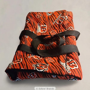 May include: A folded fabric item featuring an orange and black tiger stripe pattern with Cincinnati Bengals logos and the text Bengals. The item is secured with two black nylon straps and includes a small label reading Esfore' Brands.