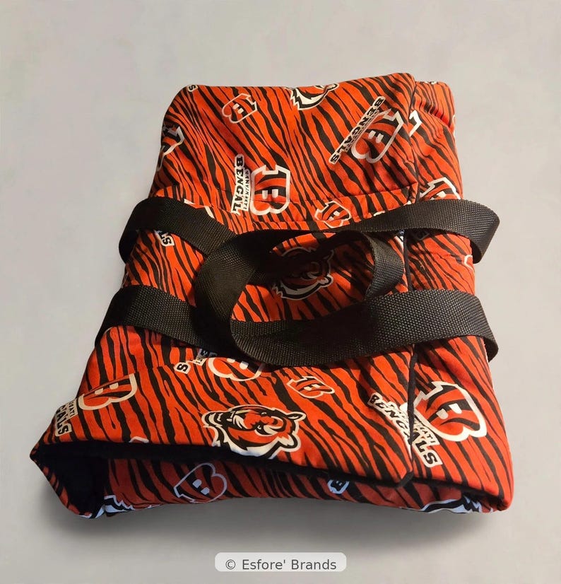 May include: A folded fabric item featuring an orange and black tiger stripe pattern with Cincinnati Bengals logos and the text Bengals. The item is secured with two black nylon straps and includes a small label reading Esfore' Brands.