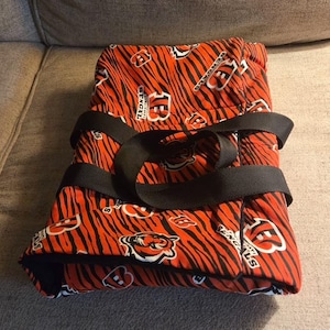May include: A folded fabric carrier featuring an orange and black tiger stripe pattern with the Cincinnati Bengals logo and text printed throughout. The item includes two black nylon webbing straps secured across the top for carrying.
