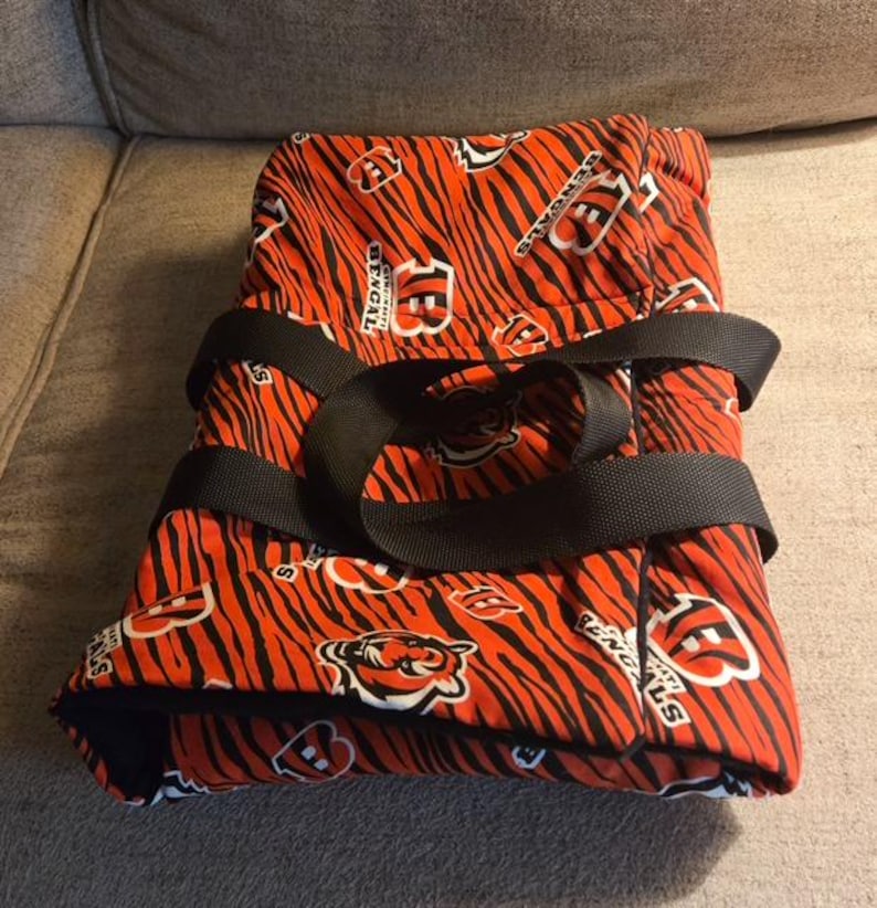 May include: A folded fabric carrier featuring an orange and black tiger stripe pattern with the Cincinnati Bengals logo and text printed throughout. The item includes two black nylon webbing straps secured across the top for carrying.