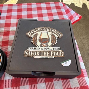 May include: A square black game board featuring a central graphic with a glass of amber liquid, wheat stalks, and the text "BOURBON & BARRELS THREE IN A ROW... THEN SAVOR THE POUR". The board has a textured surface and a small white price tag.