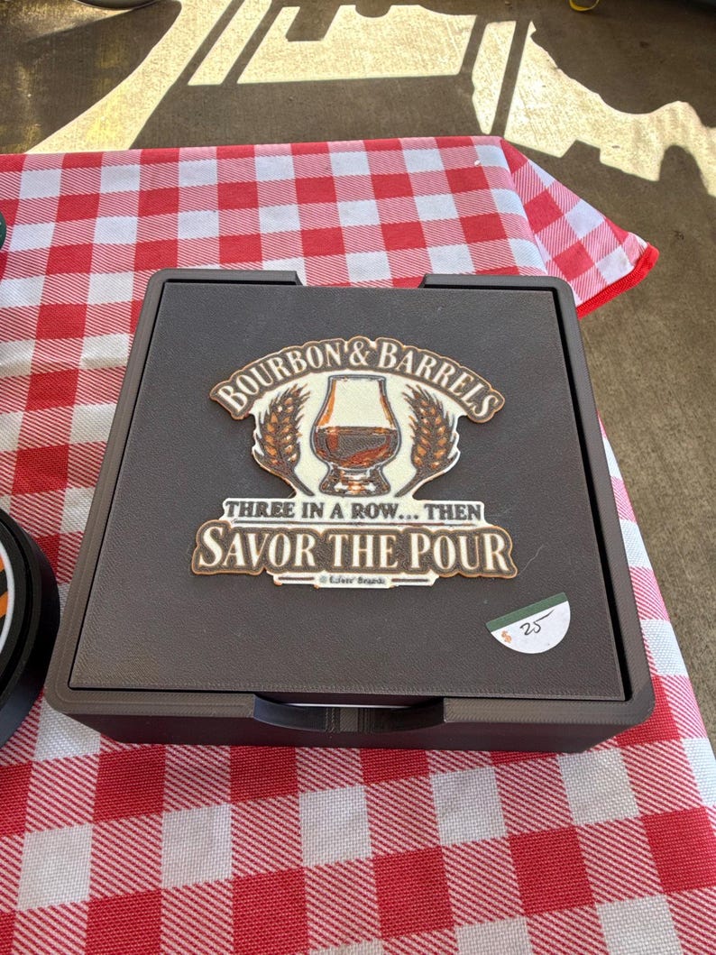 May include: A square black game board featuring a central graphic with a glass of amber liquid, wheat stalks, and the text "BOURBON & BARRELS THREE IN A ROW... THEN SAVOR THE POUR". The board has a textured surface and a small white price tag.