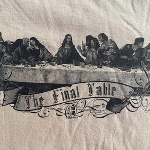 May include: A beige t-shirt featuring a black graphic print of a group of people seated at a long table. Below the table is a banner with the text The Final Table and the logo for Faded House Playing Cards brand.