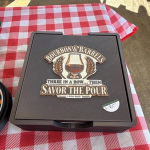 May include: A square black game board featuring a central graphic with a glass of amber liquid, wheat stalks, and the text "BOURBON & BARRELS THREE IN A ROW... THEN SAVOR THE POUR". The board has a textured surface and a small white price tag.